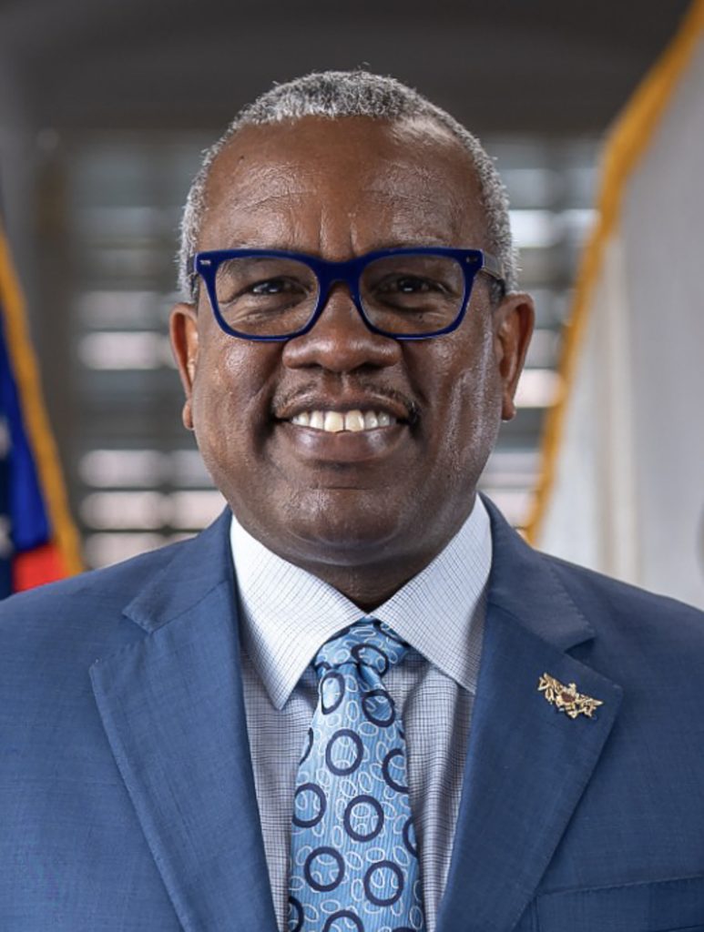 Official portrait of Governor Albert Bryan Jr., smiling, in a blue suit and blue-framed glasses, with flags behind him.